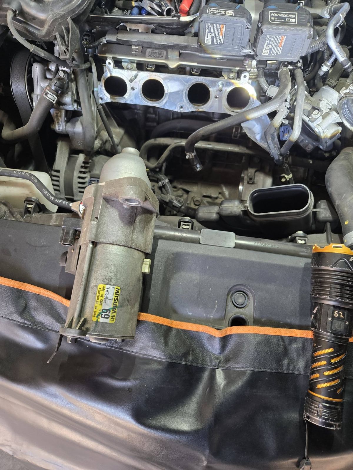 Starter motor resting on a fender cover in front of a 2012 Honda Accord engine with the intake manifold off