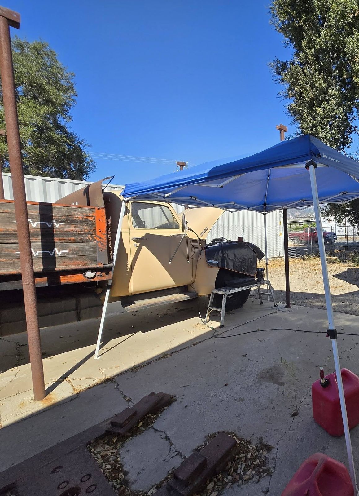 Classic flatbed truck with the hood open and a fender cover on, under a blue pop-up canopy
