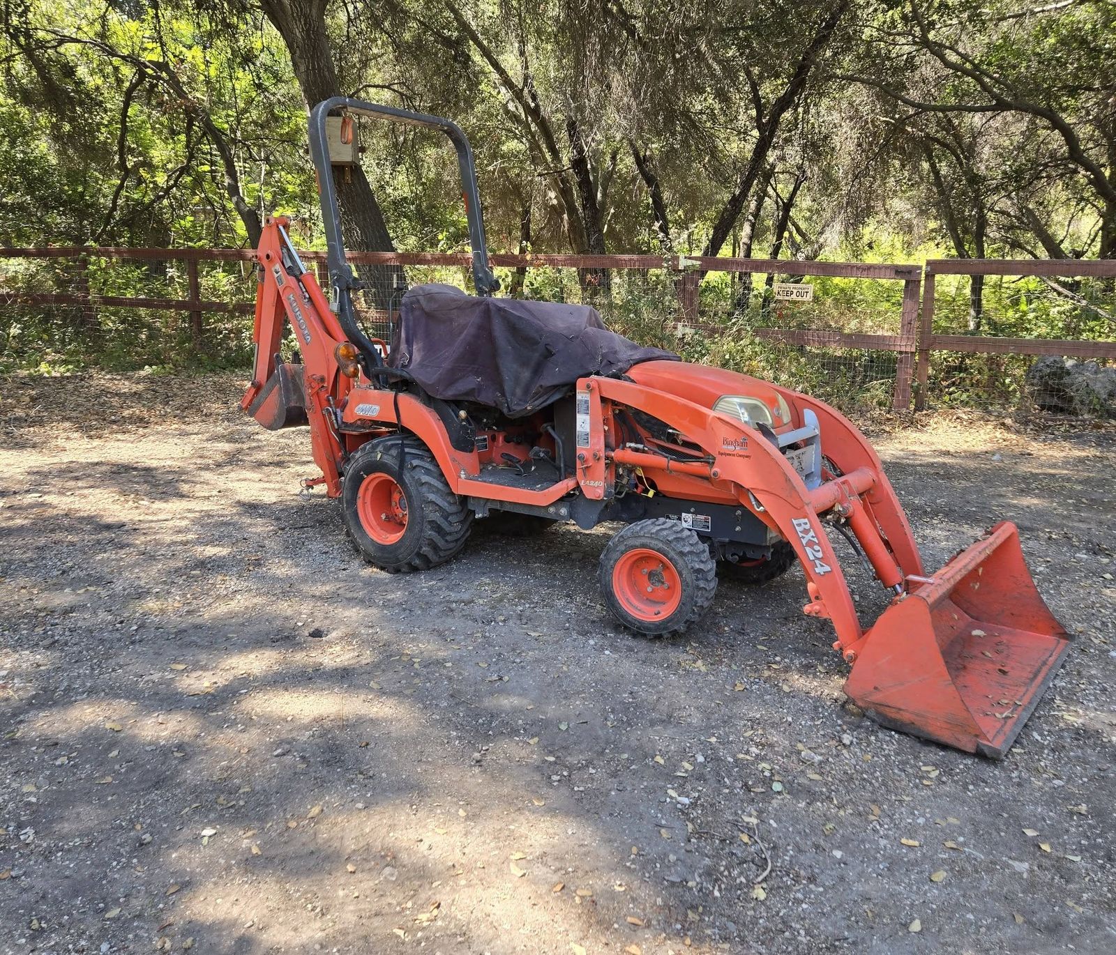 Orange BX24 compact tractor with front loader and backhoe parked under oak trees
